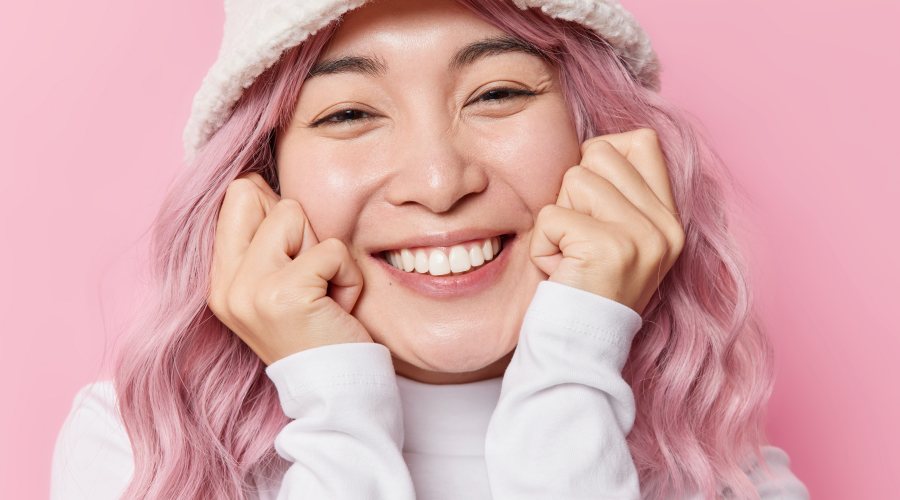 happy-sincere-asian-woman-keeps-hands-cheeks-smiles-pleasantly-shows-white-teeth-wears-fashionable-panama-white-turtleneck-feels-very-glad-isolated-pink-background-emotions-concept